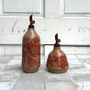 May include: Two ceramic oil and vinegar bottles with red floral designs and red tops. The bottles are on a tiled surface.