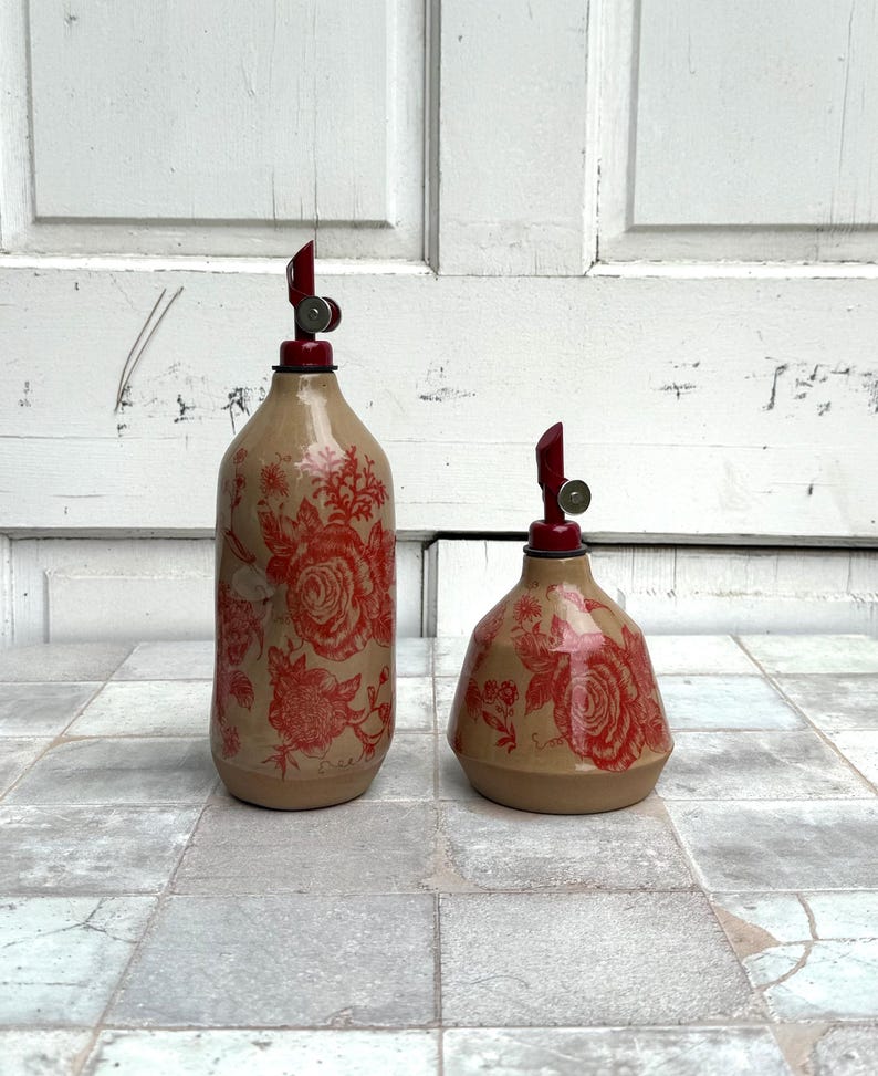 May include: Two ceramic oil and vinegar bottles with red floral designs and red tops. The bottles are on a tiled surface.