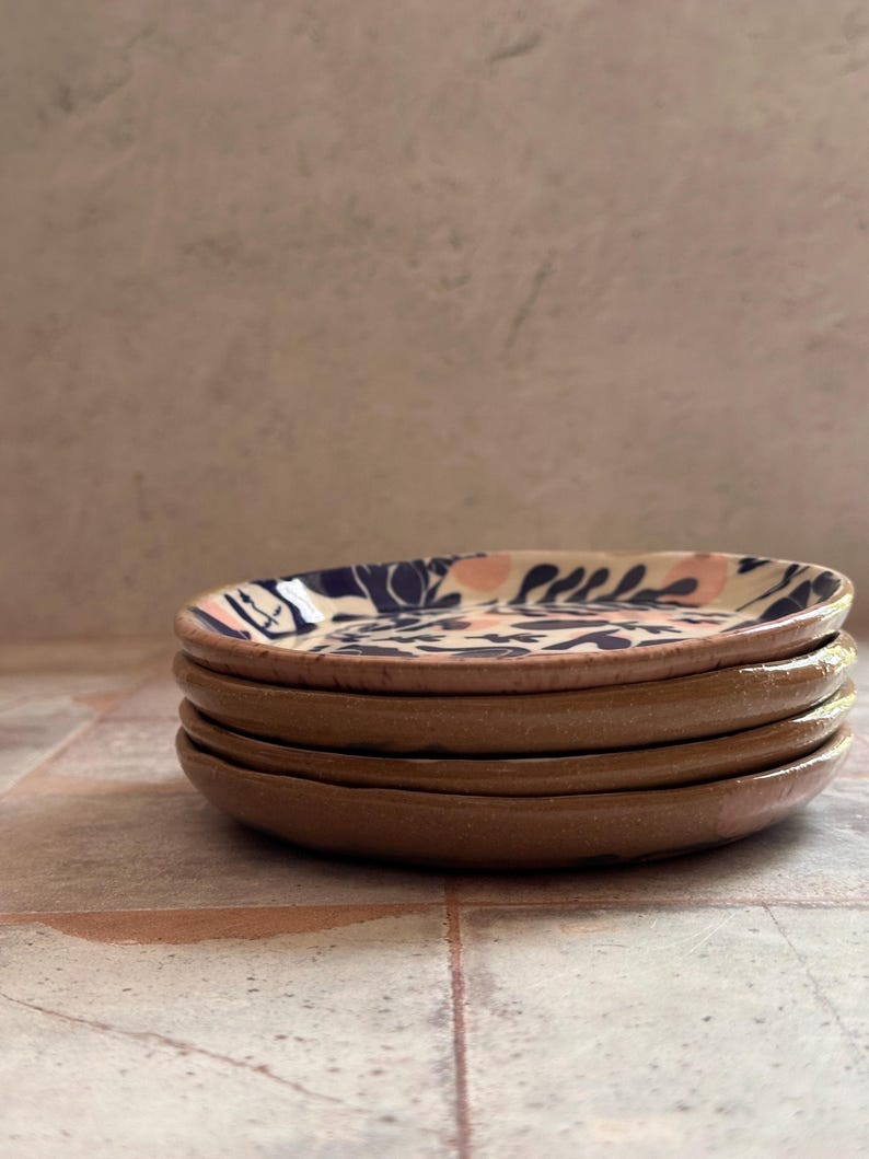 May include: A stack of four small, round ceramic plates. The plates have a brown exterior and a white interior with a blue and pink floral pattern. The plates are stacked on top of each other.