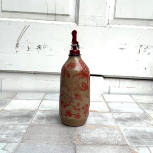 May include: A ceramic bottle with a red floral pattern and a red metal pump top. The bottle is standing on a white tiled floor.