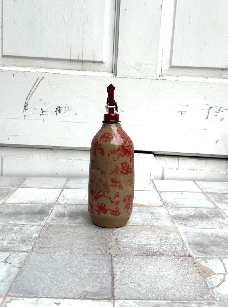 May include: A ceramic bottle with a red floral pattern and a red metal pump top. The bottle is standing on a white tiled floor.