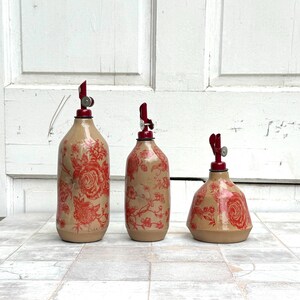 May include: Three ceramic oil and vinegar bottles with red floral patterns and red metal tops. The bottles are different sizes and shapes.
