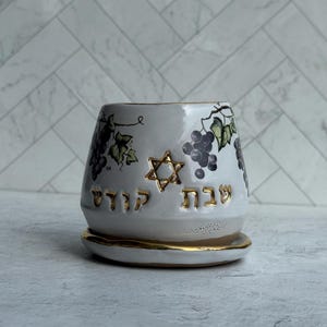 May include: White ceramic cup with a gold rim and matching saucer. The cup features a Star of David and Hebrew text in gold, along with a grape vine design in shades of purple and green. The cup is designed for religious use.