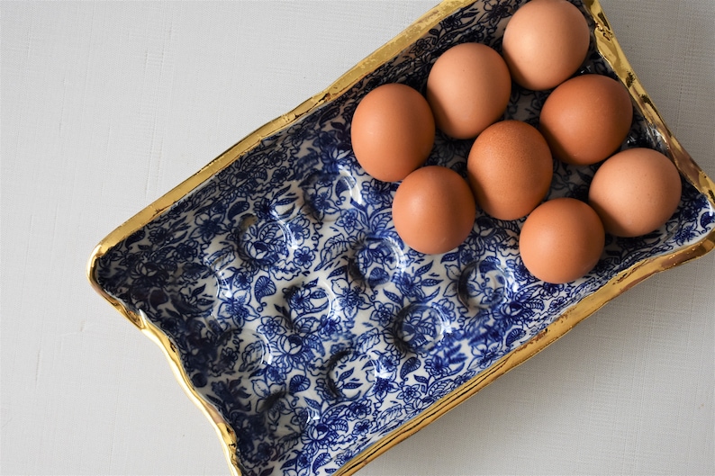 Puede incluir: Un soporte rectangular para huevos con un dise&ntilde;o floral azul y blanco y un borde dorado. Contiene ocho huevos marrones. El dise&ntilde;o recuerda a los patrones de porcelana cl&aacute;sicos. Los huevos son de color marr&oacute;n claro.