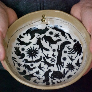 Puede incluir: Un cuenco de cer&aacute;mica con fondo blanco y dise&ntilde;os de animales y estrellas negros. El cuenco tiene una asa de metal dorado.