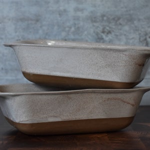 May include: Two rectangular ceramic baking dishes stacked on top of each other. The dishes have a speckled, off-white glaze on the top half and a brown base. Each dish has a handle on either side.