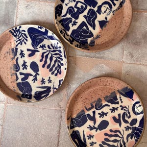 May include: Three ceramic plates with a unique design. The plates feature a cream-colored background with navy blue and pink abstract figures and botanical patterns. The rims are a speckled, earthy brown. The plates are arranged on a neutral-toned tiled surface.