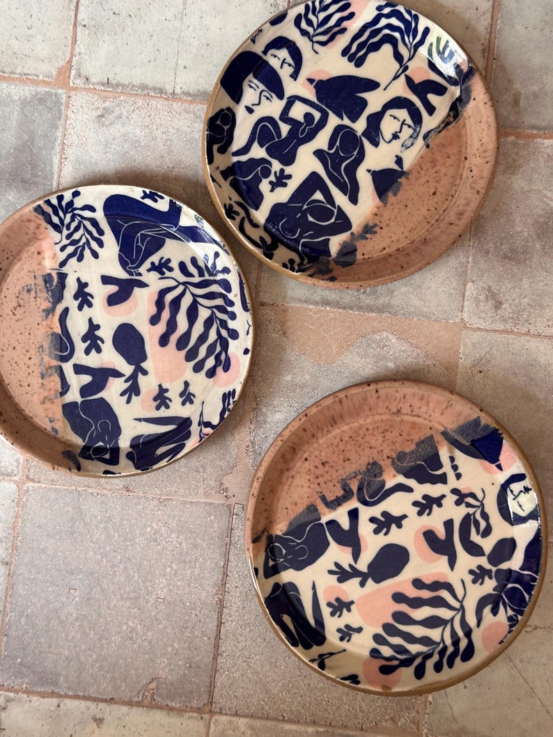 May include: Three ceramic plates with a unique design. The plates feature a cream-colored background with navy blue and pink abstract figures and botanical patterns. The rims are a speckled, earthy brown. The plates are arranged on a neutral-toned tiled surface.