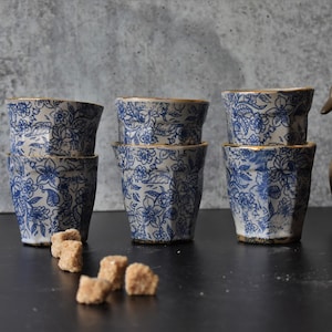 May include: Six blue and white ceramic cups with gold trim and a floral pattern. The cups are stacked in pairs and arranged on a black surface.