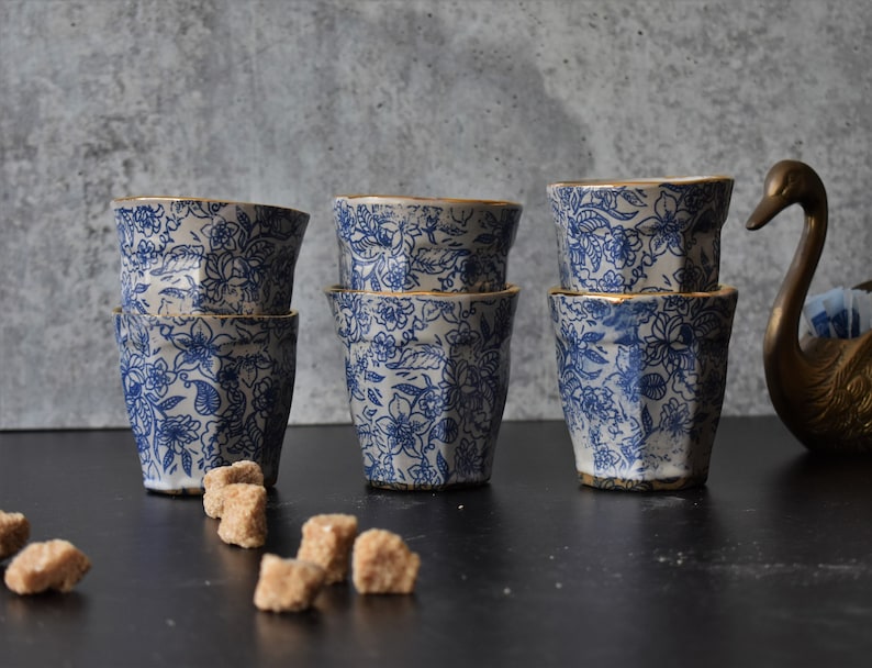 May include: Six blue and white ceramic cups with gold trim and a floral pattern. The cups are stacked in pairs and arranged on a black surface.