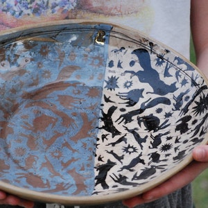May include: A decorative ceramic bowl with a split design. One side features a blue and brown pattern of animal silhouettes, while the other side has black animal silhouettes on a cream background. The bowl has a beige rim.