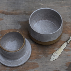 Peut inclure: Un beurrier en c&eacute;ramique grise avec un couvercle et un couteau &agrave; beurre. Le plat a une finition mouchet&eacute;e et une base marron. Le couteau &agrave; beurre a un manche d&eacute;coratif. Les articles sont sur une surface en bois.