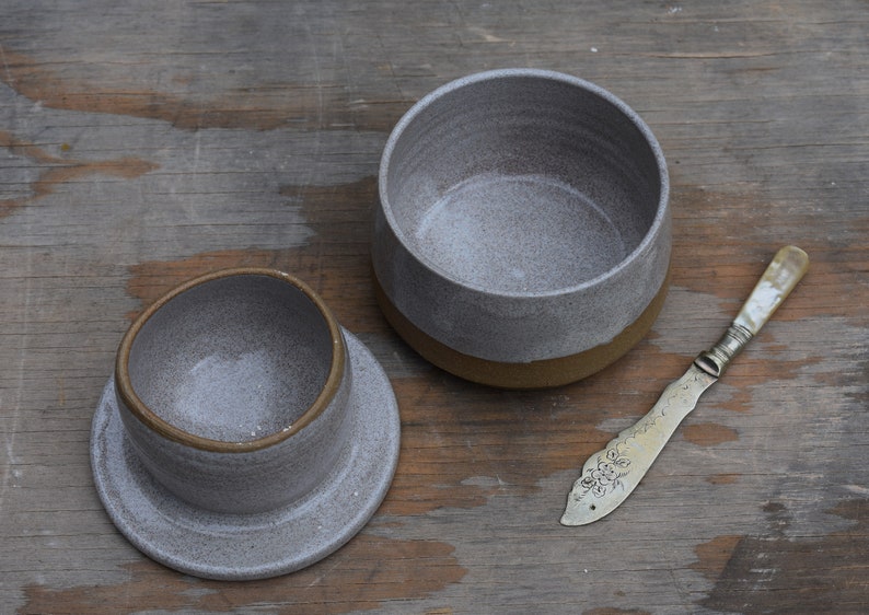 Peut inclure: Un beurrier en c&eacute;ramique grise avec un couvercle et un couteau &agrave; beurre. Le plat a une finition mouchet&eacute;e et une base marron. Le couteau &agrave; beurre a un manche d&eacute;coratif. Les articles sont sur une surface en bois.