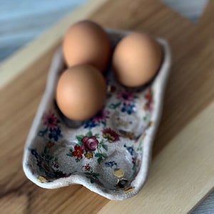 May include: A decorative egg holder with a floral design, containing three brown eggs. The holder has a cream base with a colourful floral pattern and gold detailing. The eggs are a light brown colour.