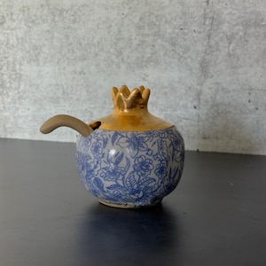 May include: A ceramic pomegranate-shaped container with a gold-colored crown and a blue floral pattern. A small, curved spoon rests on the side. The container is on a dark surface.