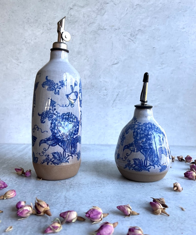 Two ceramic oil and vinegar bottles with blue and white floral designs. The bottles have a spout and a stopper. The bottles are sitting on a light gray surface with dried rose buds scattered around them.