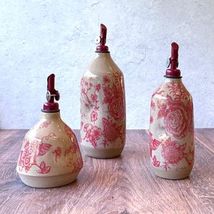 May include: Three ceramic oil and vinegar bottles with red floral designs and red pump tops. The bottles are different sizes and are sitting on a wooden surface.
