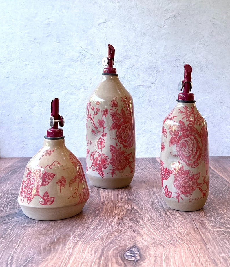 May include: Three ceramic oil and vinegar bottles with red floral designs and red pump tops. The bottles are different sizes and are sitting on a wooden surface.
