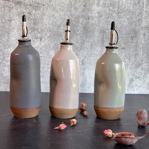 May include: Three ceramic oil and vinegar bottles with a speckled glaze finish. The bottles have a brown rim and a metal pour spout with a black top. The bottles are arranged in a row on a black surface.