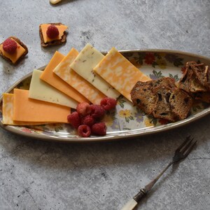 K&ouml;nnte beinhalten: Eine wei&szlig;e ovale Platte mit Blumenmuster enth&auml;lt eine Auswahl an K&auml;se, Crackern und Himbeeren. Die Platte befindet sich auf einer grauen Oberfl&auml;che.