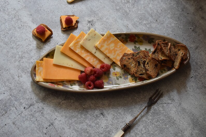 K&ouml;nnte beinhalten: Eine wei&szlig;e ovale Platte mit Blumenmuster enth&auml;lt eine Auswahl an K&auml;se, Crackern und Himbeeren. Die Platte befindet sich auf einer grauen Oberfl&auml;che.