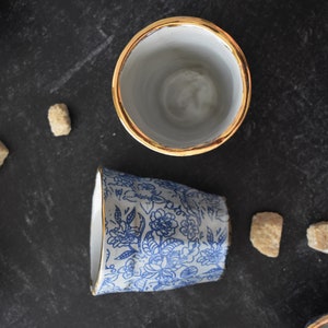 May include: Two ceramic mugs with gold rims. One mug is white with a blue floral pattern, the other is plain white.
