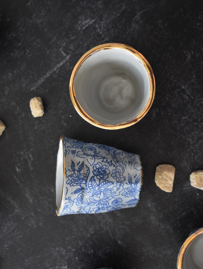May include: Two ceramic mugs with gold rims. One mug is white with a blue floral pattern, the other is plain white.