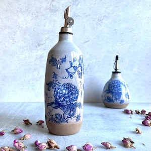 Two ceramic oil and vinegar bottles with blue and white floral designs. The larger bottle has a silver pour spout. The bottles are sitting on a light gray surface with dried pink rose buds scattered around them.