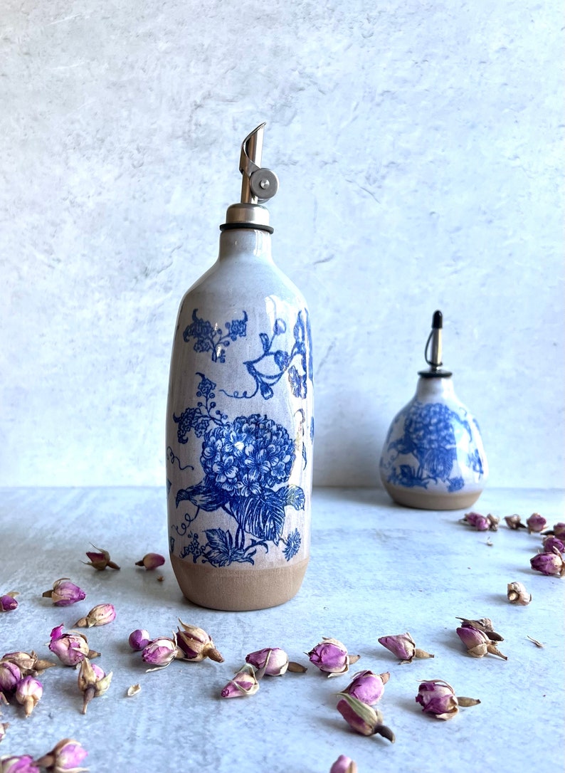 Two ceramic oil and vinegar bottles with blue and white floral designs. The larger bottle has a silver pour spout. The bottles are sitting on a light gray surface with dried pink rose buds scattered around them.