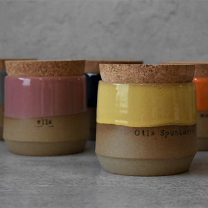 May include: Five ceramic jars with cork lids. The jars are in different colors: gray, pink, blue, yellow, and orange. Each jar has a different name written on the side: "Ella", "Otis Spunkmeyer", "Remarkable", and two others that are not visible.