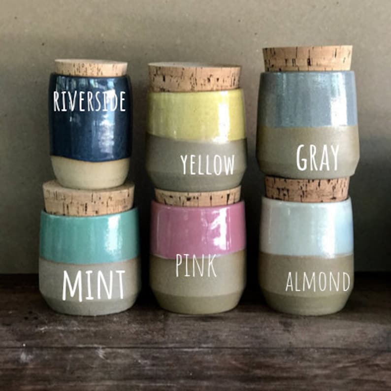 Op de afbeelding: Zes kleine keramische potjes met kurken stoppen, elk in een andere kleur: blauw, geel, grijs, mint, roze en amandel. De kleurnamen staan in witte tekst op de potjes. Geschikt voor het bewaren van kleine items.