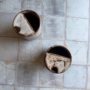 May include: Two handmade ceramic containers with a speckled beige and brown glaze. Each cylindrical vessel has a diagonal cut and a small hole. The interior is dark brown.