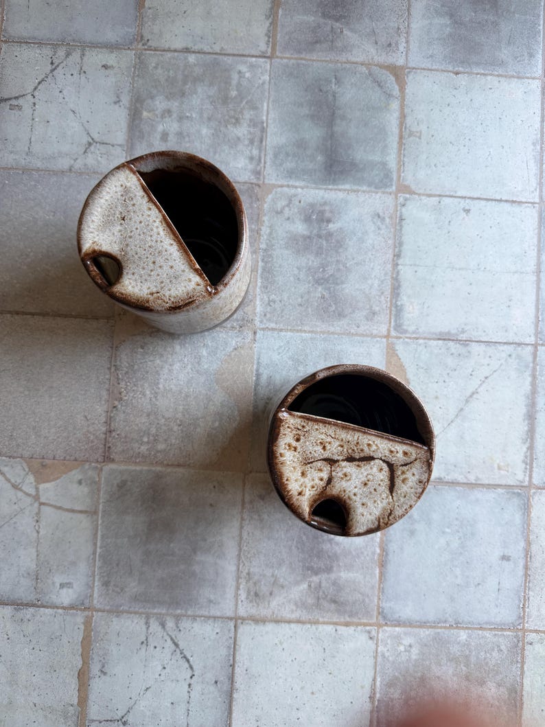 May include: Two handmade ceramic containers with a speckled beige and brown glaze. Each cylindrical vessel has a diagonal cut and a small hole. The interior is dark brown.