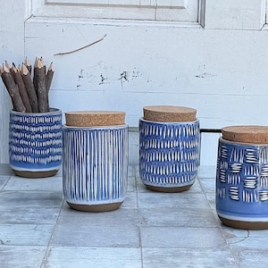 May include: Four blue ceramic jars with cork lids and various textured patterns. One jar holds pencils. The jars are in shades of blue and white, with a natural cork color. The jars are on a tiled surface.