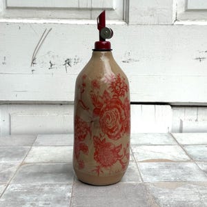 May include: A ceramic bottle with a red floral design and a red and silver pour spout. The bottle is sitting on a tiled floor.