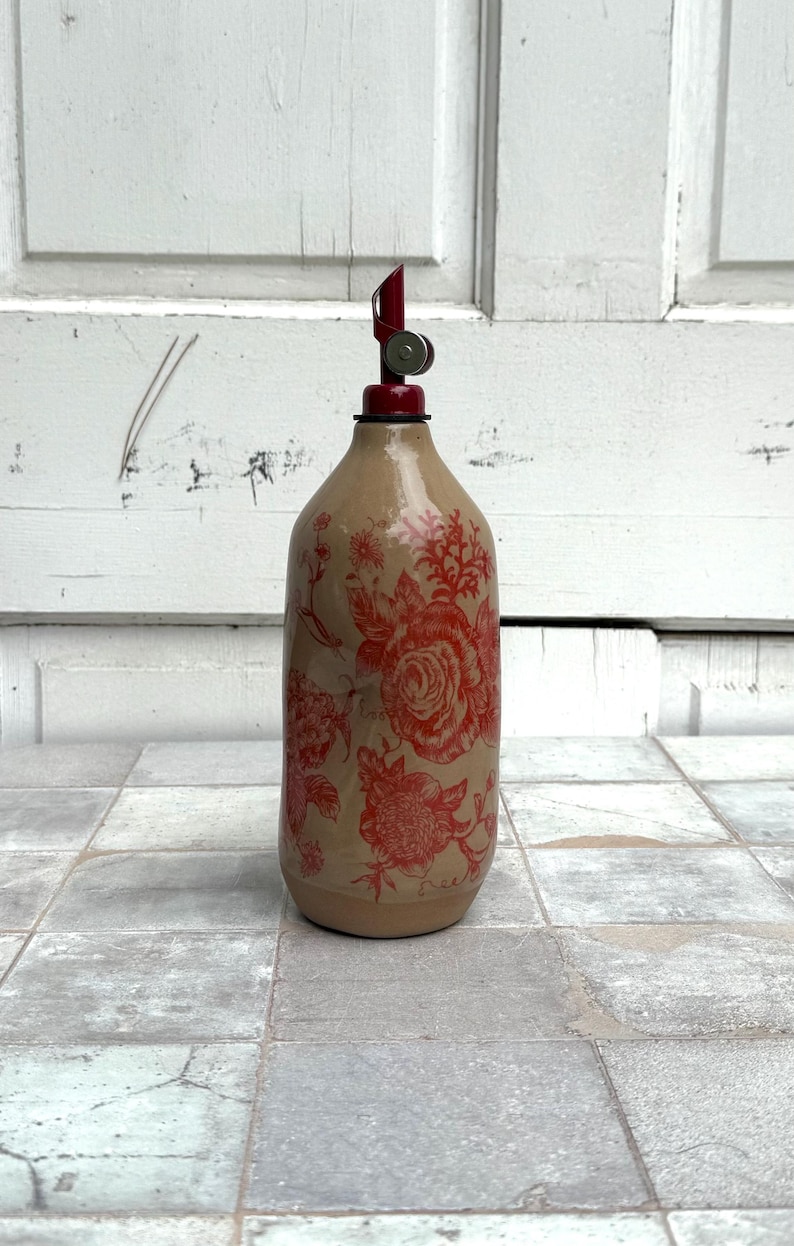 May include: A ceramic bottle with a red floral design and a red and silver pour spout. The bottle is sitting on a tiled floor.