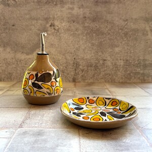 May include: A ceramic oil bottle and small dish set. The bottle has a brown base and a tan upper section with a silver spout. Both pieces are decorated with a repeating pattern of yellow, orange, and black shapes on a white background.