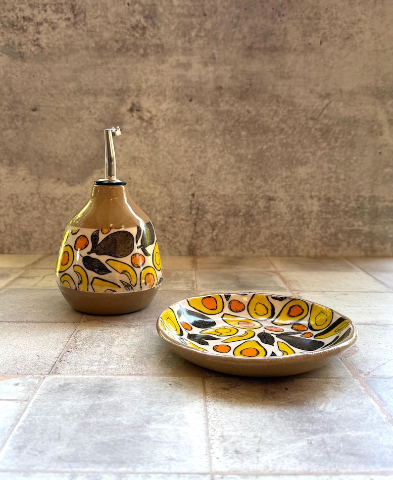 May include: A ceramic oil bottle and small dish set. The bottle has a brown base and a tan upper section with a silver spout. Both pieces are decorated with a repeating pattern of yellow, orange, and black shapes on a white background.
