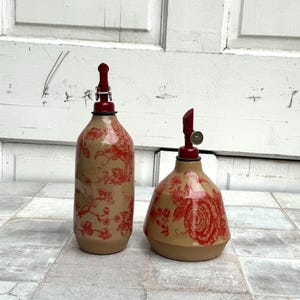 May include: Two ceramic oil and vinegar bottles with red floral designs and red metal tops. The bottles are on a tiled surface.