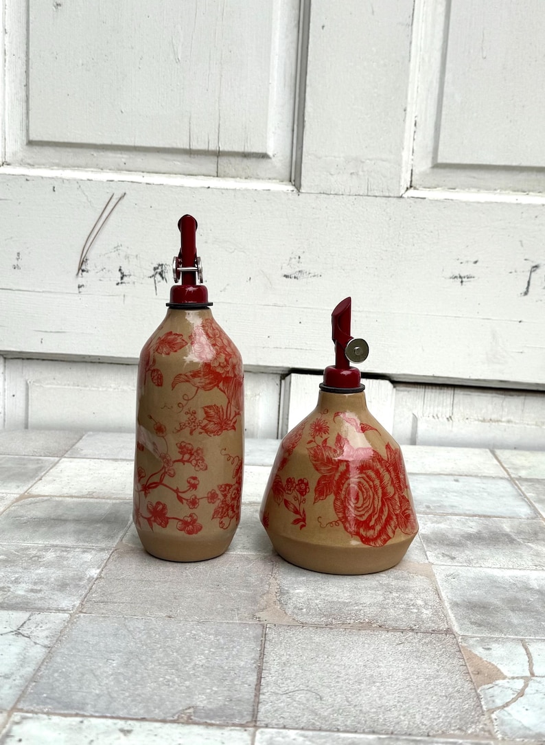 May include: Two ceramic oil and vinegar bottles with red floral designs and red metal tops. The bottles are on a tiled surface.