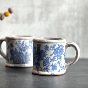 May include: Two ceramic mugs with blue floral patterns. The mugs have a cream-colored base and a rounded handle. The floral design is a classic blue and white pattern. The mugs are on a dark surface.