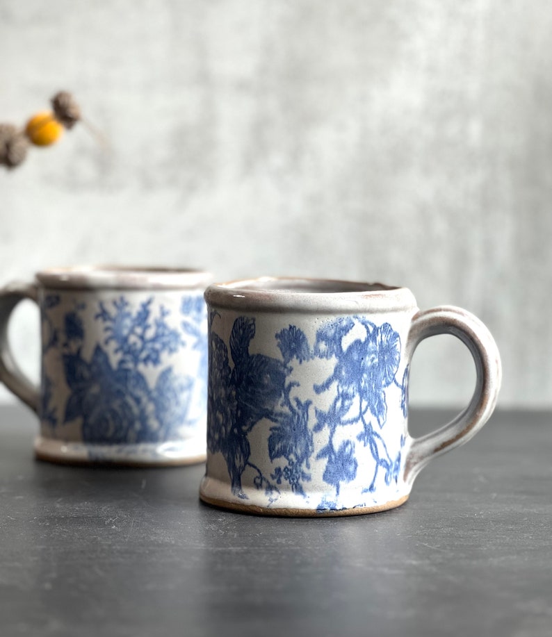 May include: Two ceramic mugs with blue floral patterns. The mugs have a cream-colored base and a rounded handle. The floral design is a classic blue and white pattern. The mugs are on a dark surface.