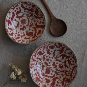 May include: Two white ceramic bowls with red and white animal silhouettes. The bowls have a repeating pattern of foxes, rabbits, birds, and stars. The bowls are on a gray surface.
