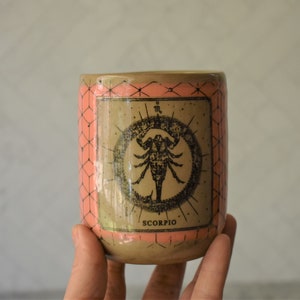 May include: A ceramic mug with a pink and brown geometric pattern. The mug features a black and white illustration of a scorpion with the word "SCORPIO" printed below it.