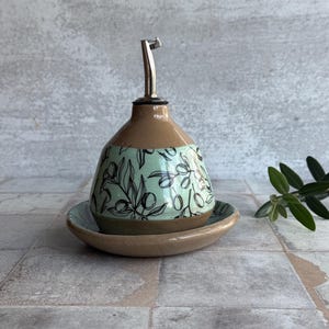 May include: A ceramic olive oil dispenser with a matching saucer. The dispenser has a brown base and a light green section with a black olive branch design. It features a silver spout and is designed for kitchen use.