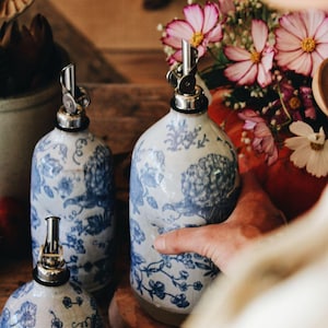 May include: Three ceramic oil dispensers featuring a white base with blue floral patterns and silver-toned metal pour spouts. The dispensers have rounded bodies and are arranged together, showcasing a consistent design across all three pieces.