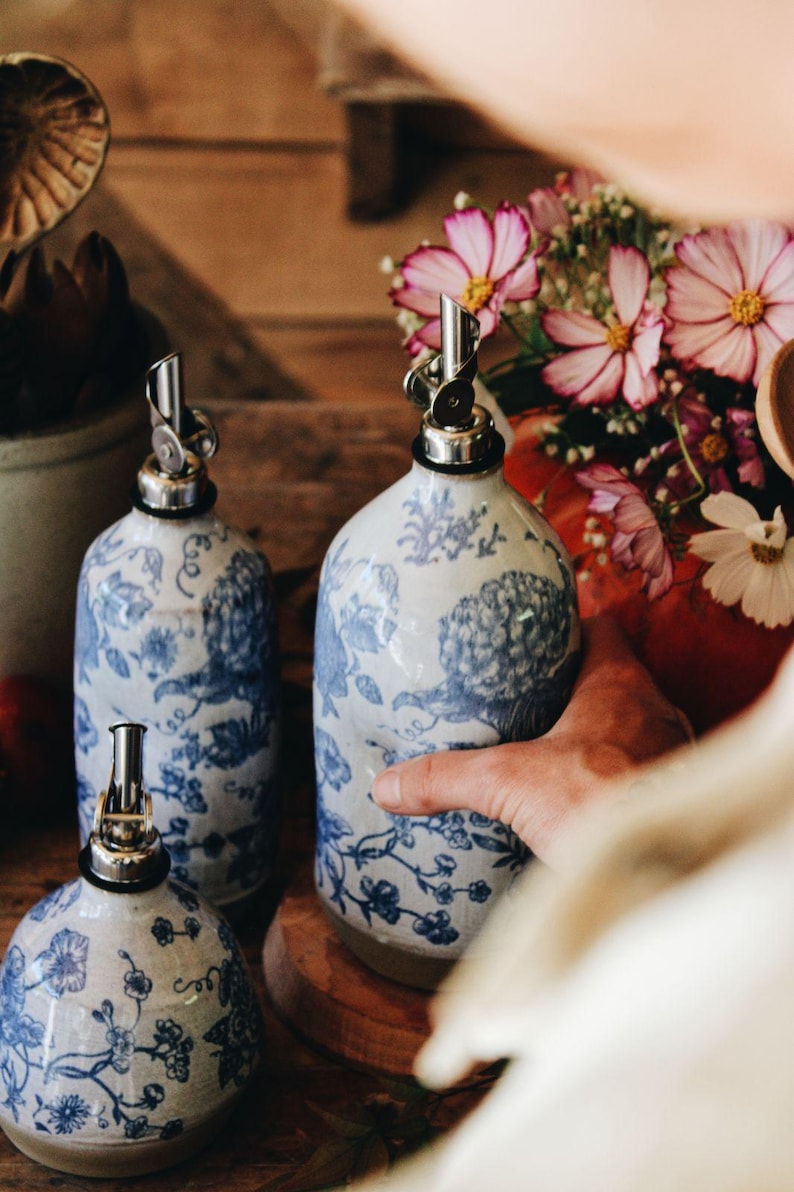 May include: Three ceramic oil dispensers featuring a white base with blue floral patterns and silver-toned metal pour spouts. The dispensers have rounded bodies and are arranged together, showcasing a consistent design across all three pieces.