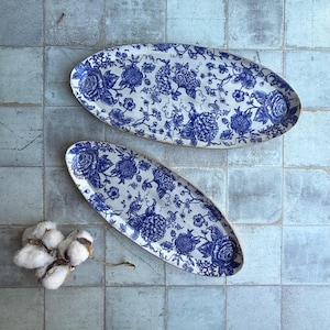 May include: Two oval serving platters with a blue floral pattern on a white background. The platters have a classic design with roses and other flowers. The platters are on a blue tiled surface.