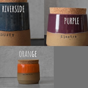 Op de afbeelding: Drie keramische potten met kurken deksels. E&eacute;n pot is marineblauw met de woorden "Riverside" en "Dusty" erop gedrukt. Een andere is paars met "Purple" en "Electra". De derde is oranje met "Orange" en "marmalade".