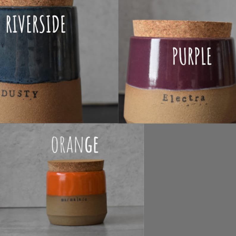 Op de afbeelding: Drie keramische potten met kurken deksels. E&eacute;n pot is marineblauw met de woorden "Riverside" en "Dusty" erop gedrukt. Een andere is paars met "Purple" en "Electra". De derde is oranje met "Orange" en "marmalade".